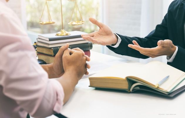 Zwei Personen in einer Rechtsberatung, die über einen Schreibtisch mit Gesetzbüchern und Justitia-Waagen gestikulieren.