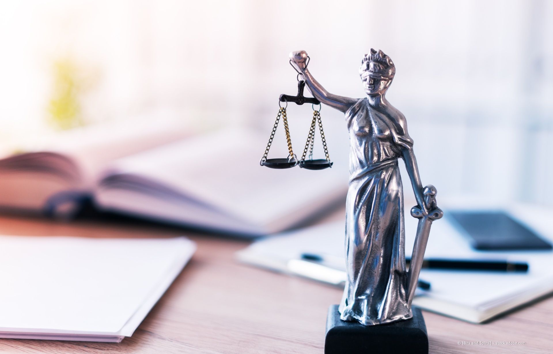 Eine Justitia-Statue steht auf einem Schreibtisch; im Hintergrund sind ein offenes Buch und Papiere zu sehen.