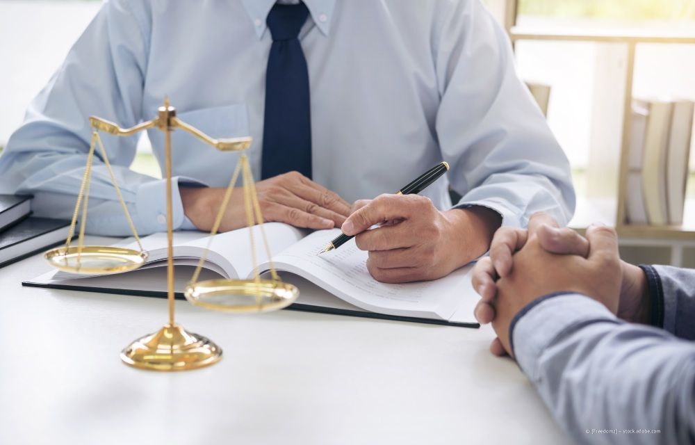 Anwalt mit Krawatte prüft Dokumente an einem Schreibtisch, im Vordergrund eine Messingwaage der Justitia.