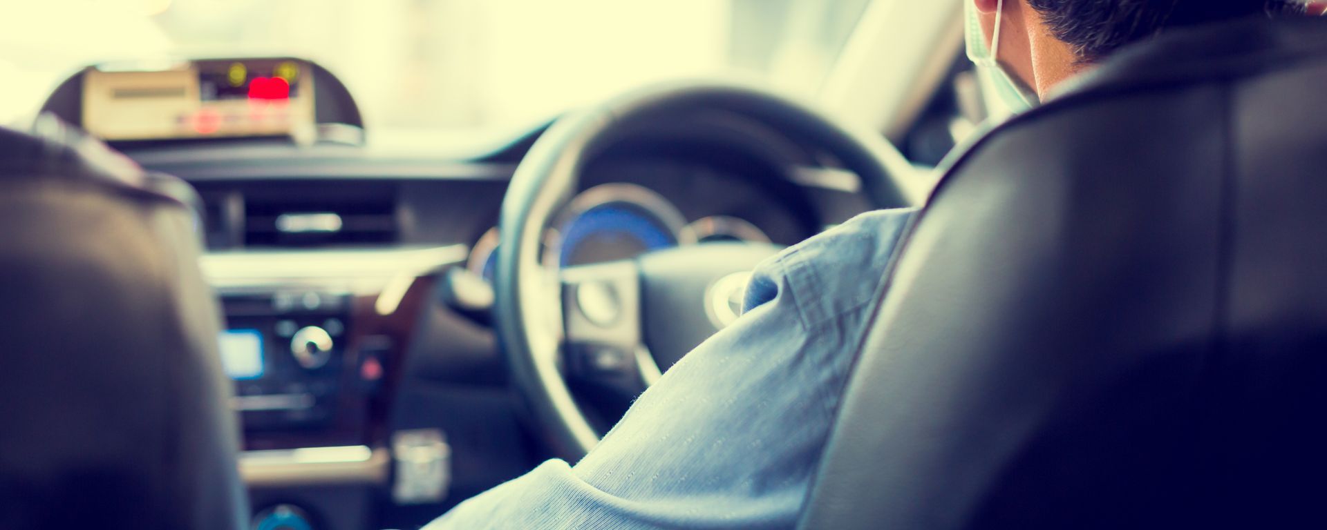 Une personne conduisant une voiture, vue depuis la banquette arrière. Le volant et le tableau de bord sont visibles.