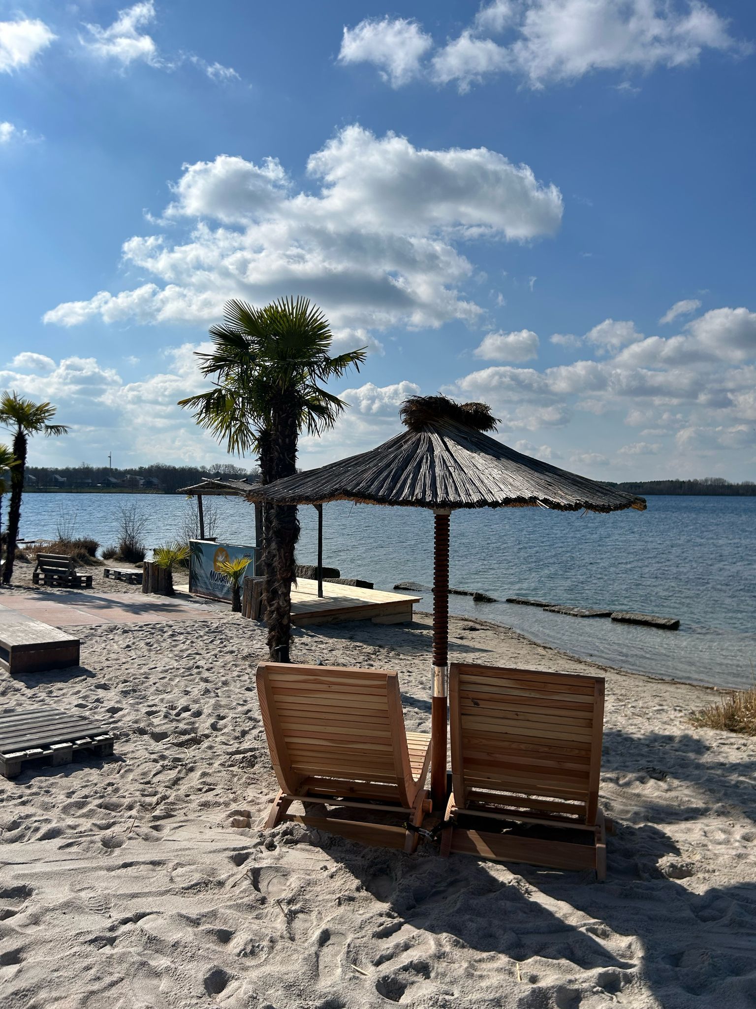 Geschwungene Liegestühle aus Holz auf einer Terrasse mit einem Sonnenschirm aus Stroh an einem See mit Strand