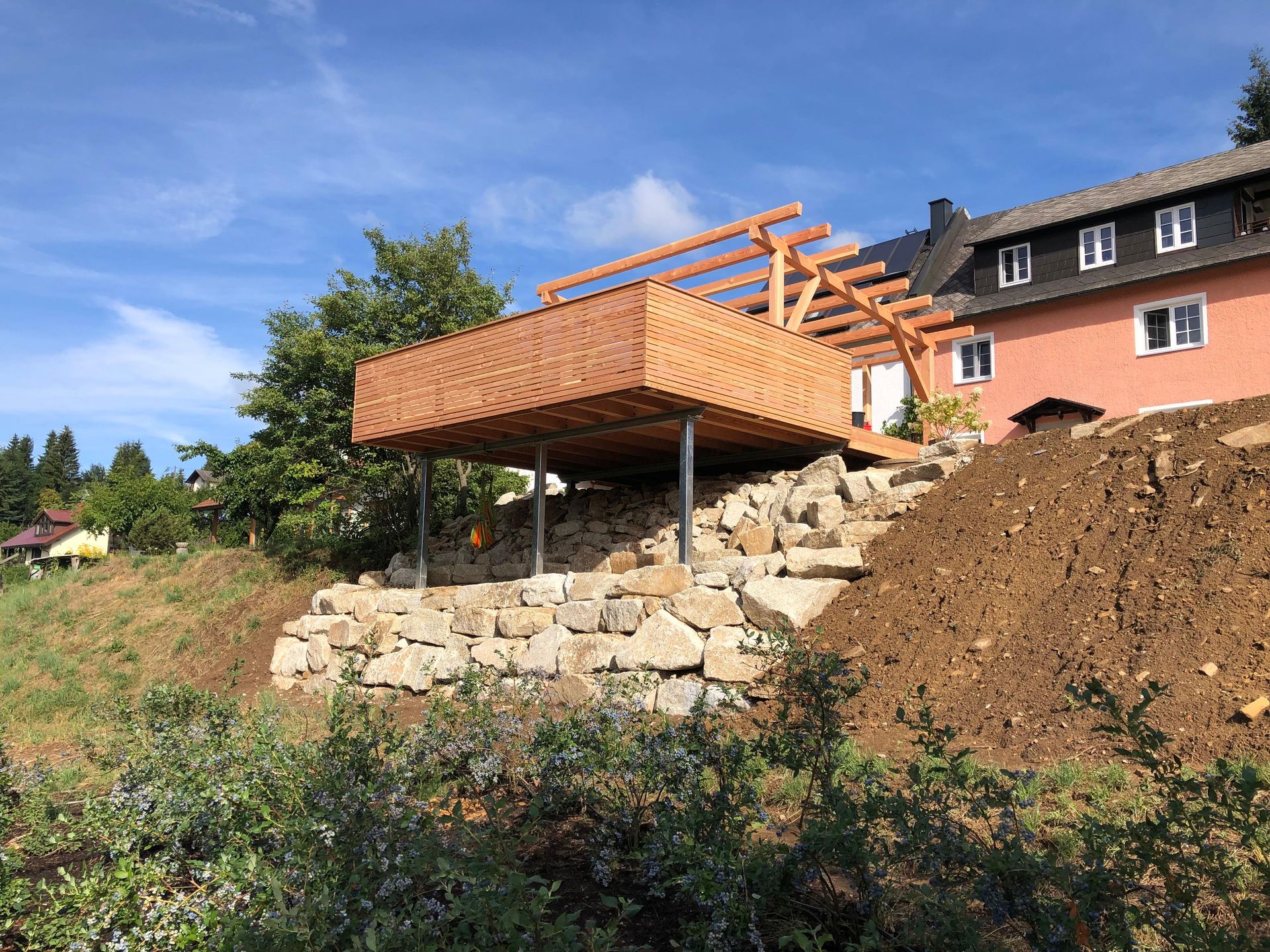Holzterrasse mit Pergola und Balkon an einem Hang, der zur Befestigung mit Natursteinen versehen ist