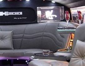 Interior de una limusina: asientos de cuero gris, copas de champán, una pequeña mesa y una pantalla de televisión con un logotipo.