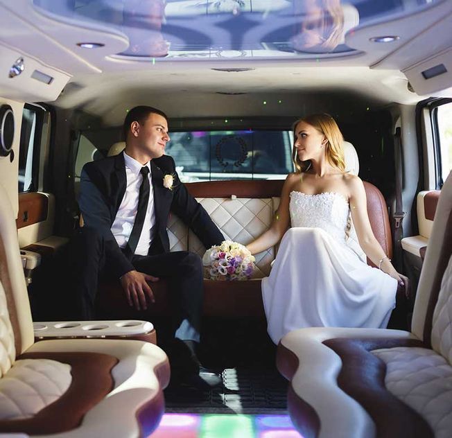 Los novios están sentados en una limusina, mirándose, sosteniendo flores. Interior con asientos de cuero e iluminación cenital.