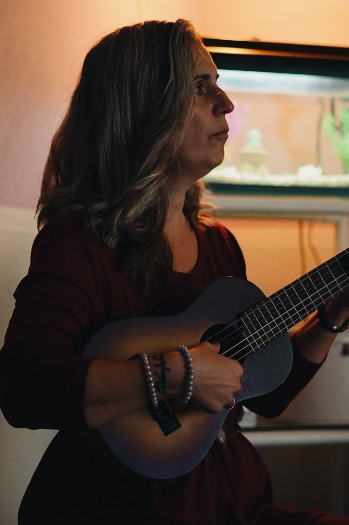 Mujer tocando una guitarra pequeña en un interior. Lleva una blusa granate y pulseras.