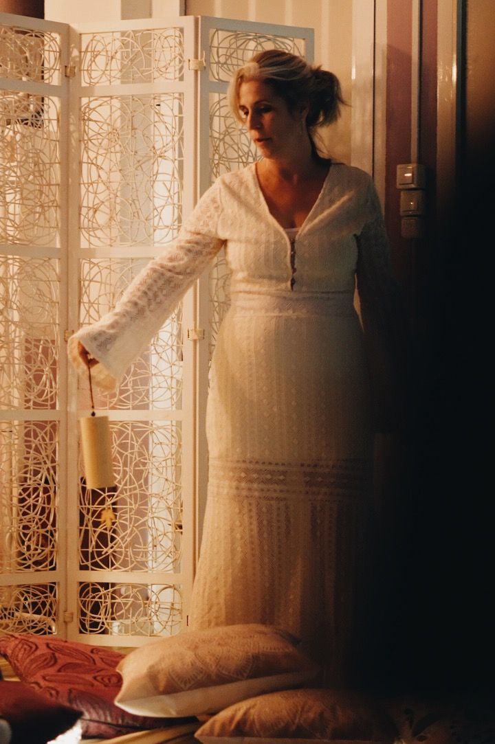 Mujer con vestido blanco, sosteniendo un bolso, de pie junto a un biombo ornamentado, con almohadas en primer plano.