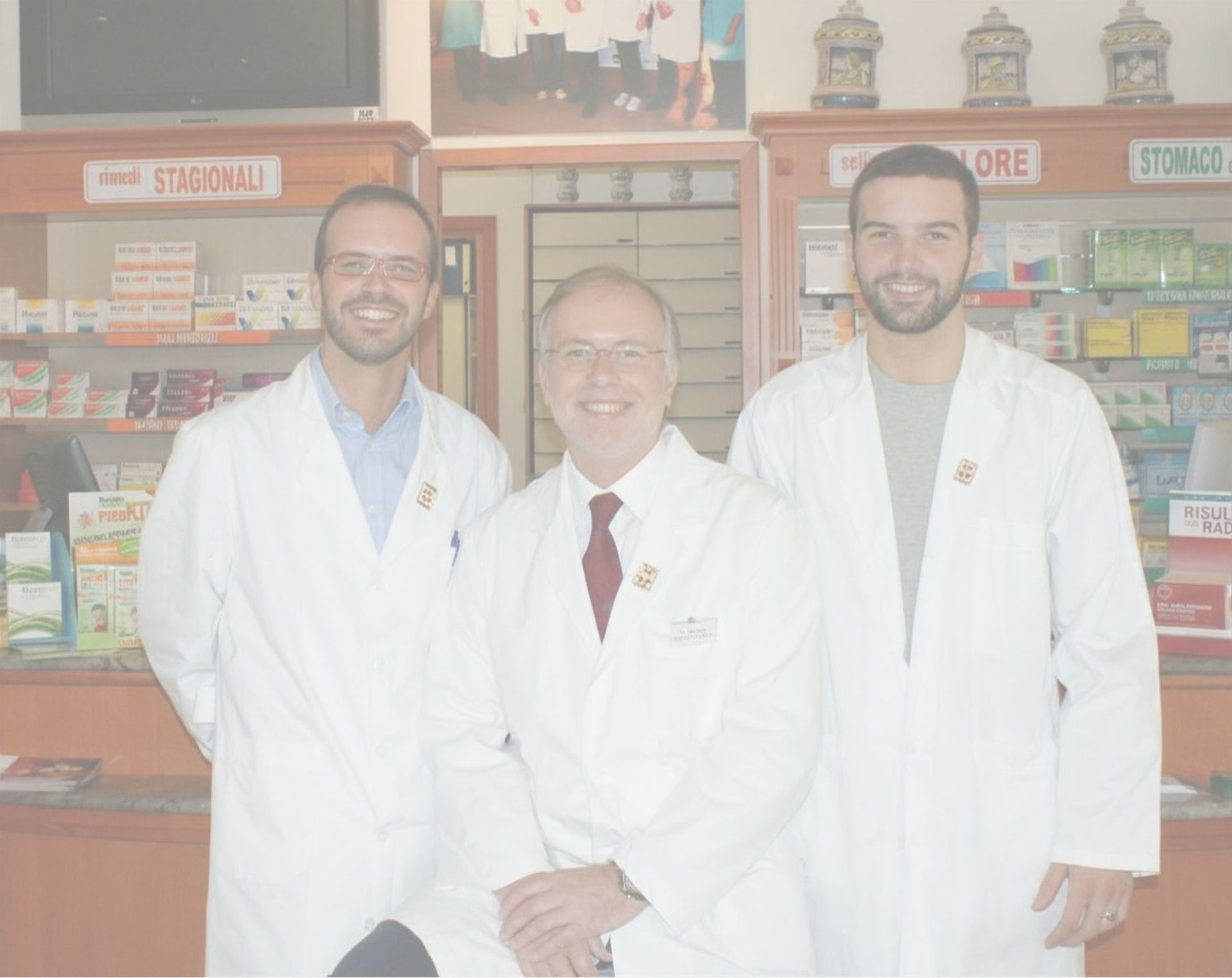 Tre persone in camice bianco in piedi in una farmacia o in un negozio, sorridenti dietro un bancone.