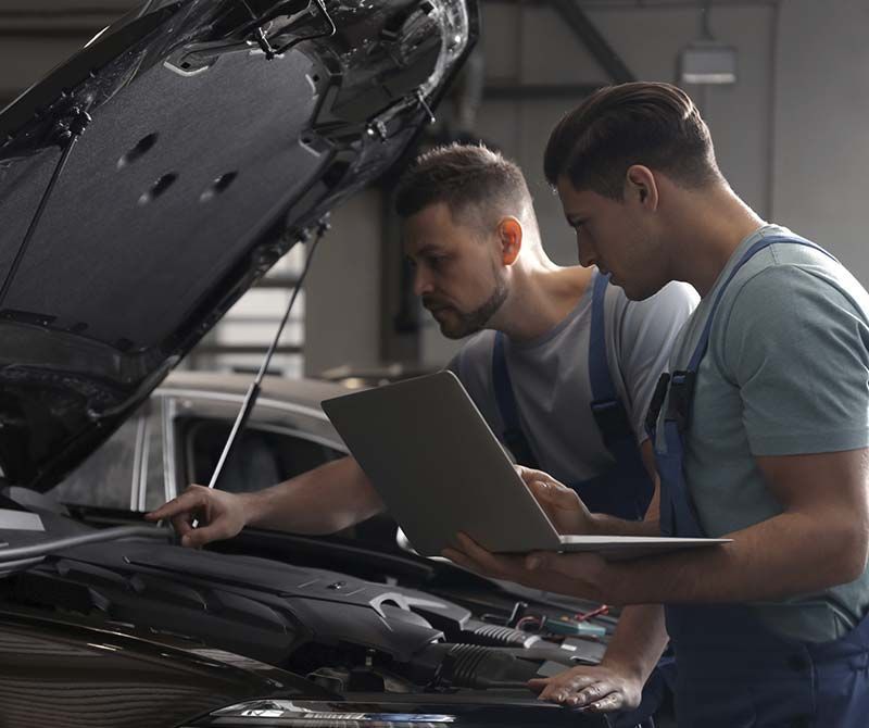 Dos hombres están trabajando en un automóvil con el capó abierto y mirando una computadora portátil.