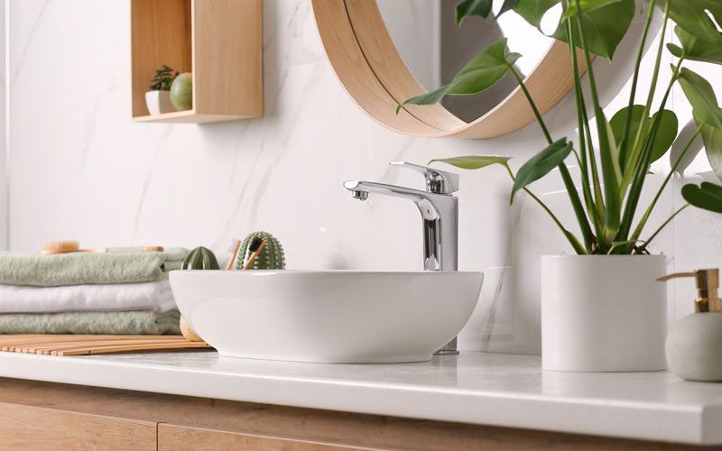 Badezimmer mit weißem Waschbecken, verchromter Armatur, Handtüchern, Spiegel, Pflanzen und Holzregal.