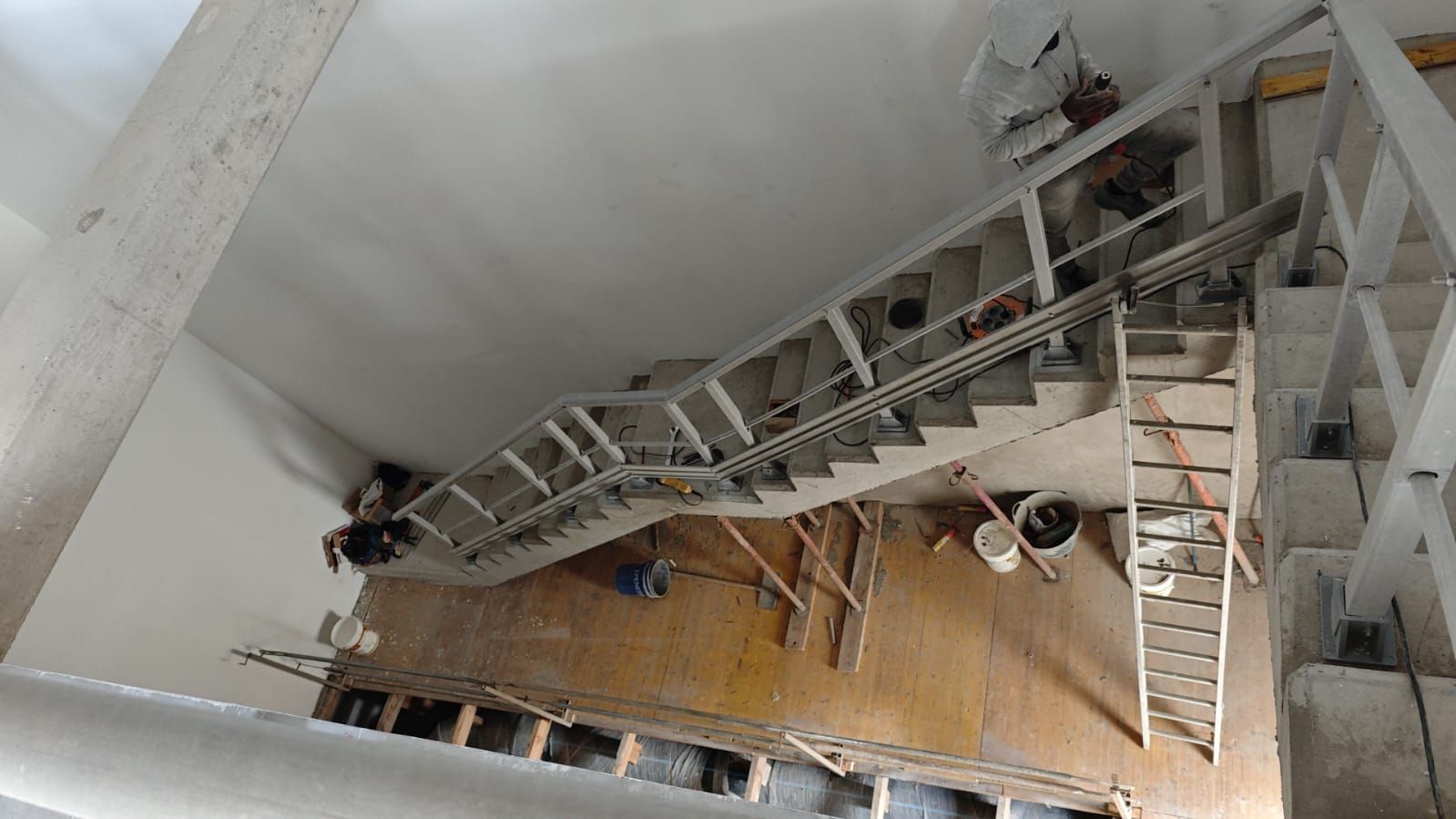 Escalera en construcción, vista desde arriba. Hay obreros presentes, junto con materiales de construcción. Paredes y techo blancos.