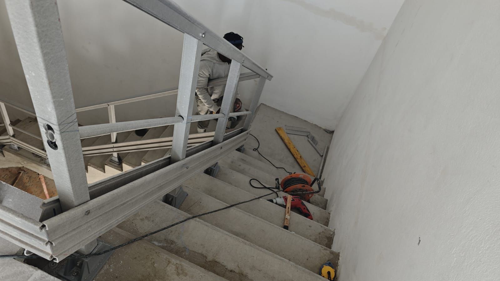 Una persona trabaja en la construcción de una escalera, con una estructura metálica y herramientas en los escalones. El espacio es un interior con paredes blancas.