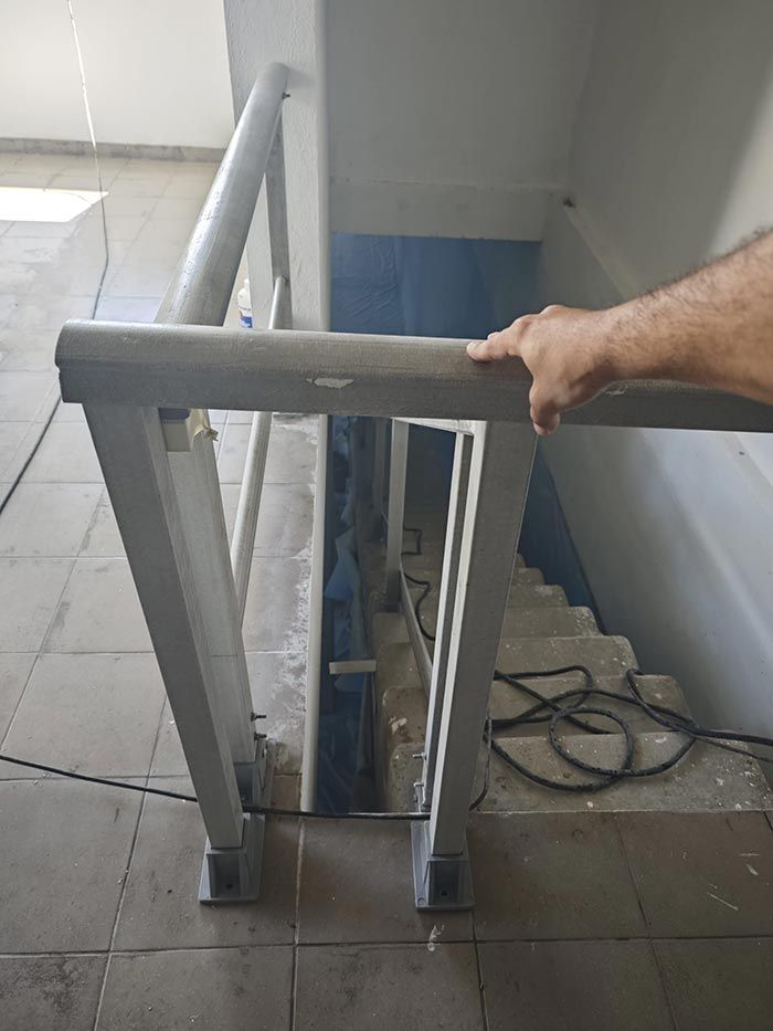 La mano de una persona se apoya en un pasamanos gris con vistas a una escalera. Las escaleras descienden a un nivel inferior; los cables eléctricos descansan en el suelo.