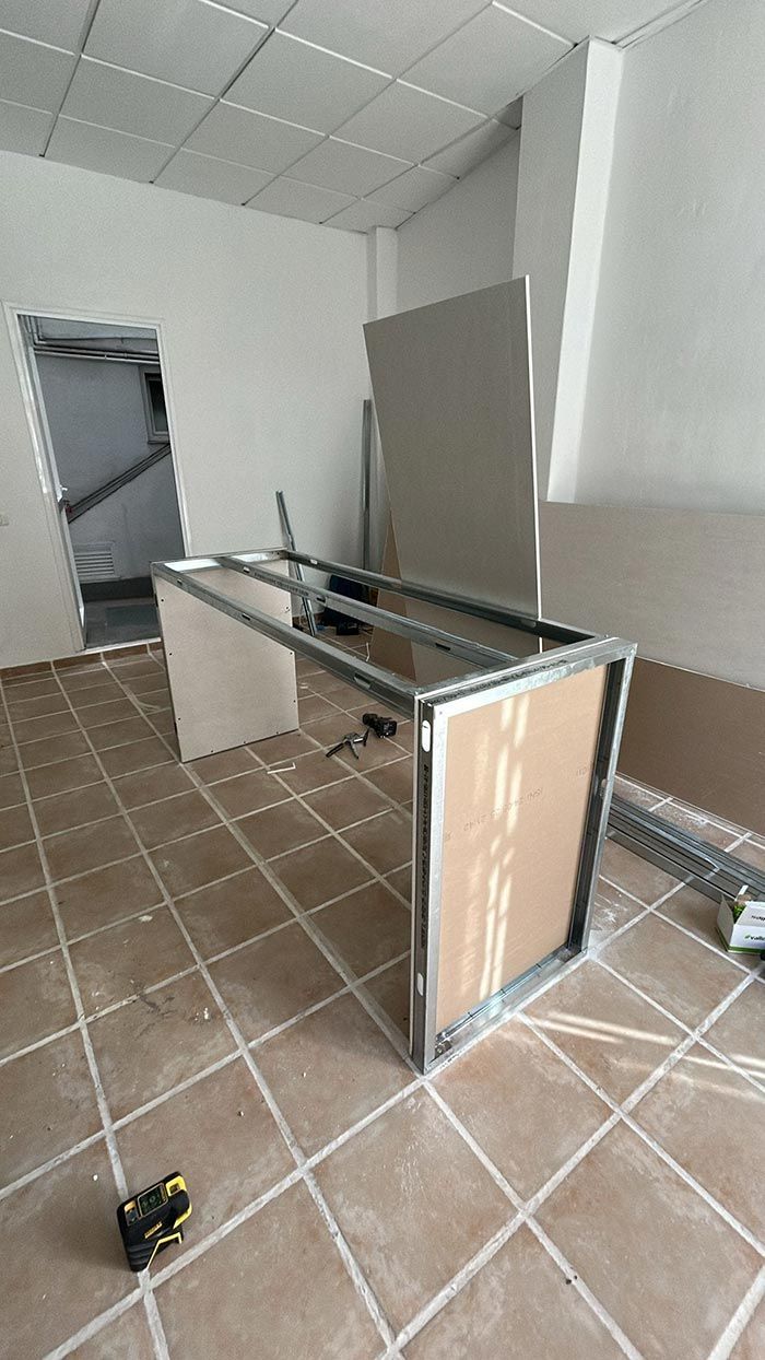 Obra interior con un escritorio y paredes parcialmente construidos, con suelo de baldosas. Se ven paneles de yeso y estructura metálica.