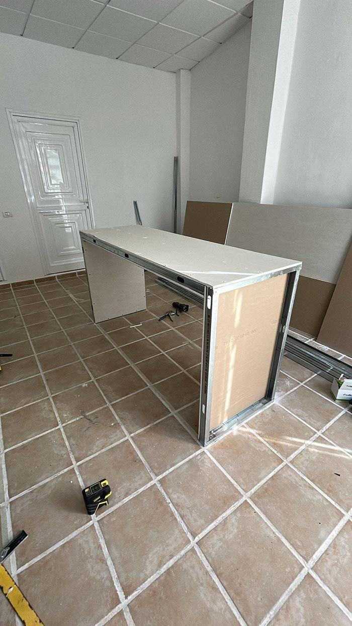 Un escritorio parcialmente construido, hecho con estructura de metal y paneles de yeso, se encuentra en una habitación con piso de baldosas y paredes sin terminar.