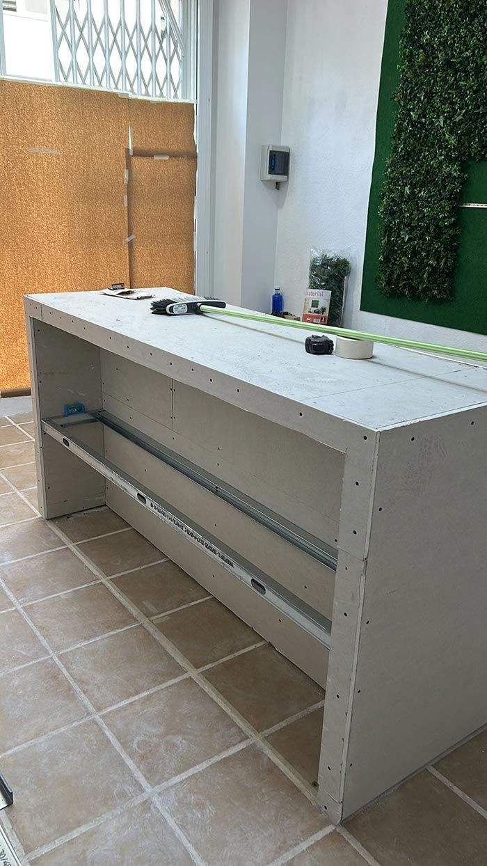 Un bar rectangular, a medio construir, con estanterías metálicas se encuentra en un interior. Encima hay herramientas y una cinta métrica.