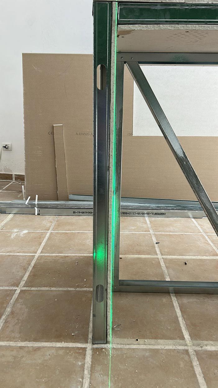 Una línea láser verde se proyecta verticalmente a lo largo de una estructura metálica. Al fondo se ven una placa de yeso y materiales de construcción.