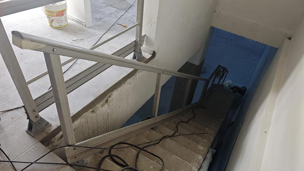 Escalera en construcción. Pasamanos metálicos claros y escalones de hormigón sin terminar conducen hacia abajo; pared azul a lo lejos.