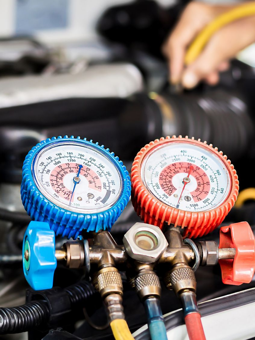 Gros plan sur les manomètres de la climatisation d'une voiture, bleus et rouges, reliés à des tuyaux, avec la main d'un mécanicien en arrière-plan.