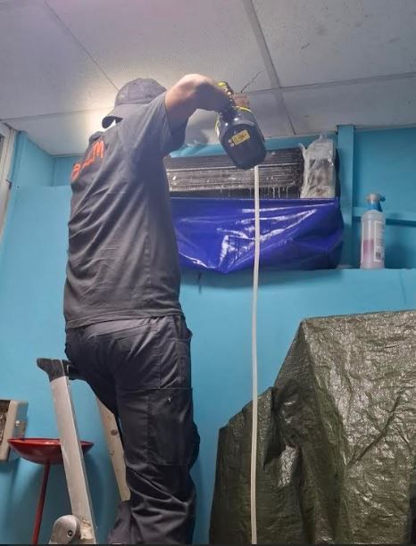 Une personne nettoyant un climatiseur. Elle se tient sur une échelle.