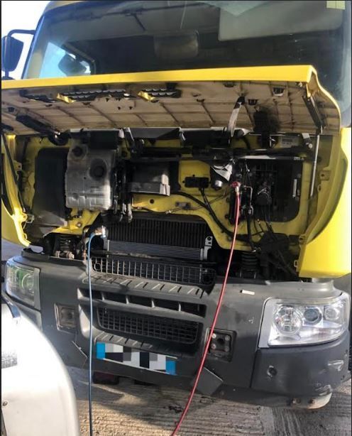 Camion jaune, capot ouvert, moteur visible, câble rouge, vue de face.