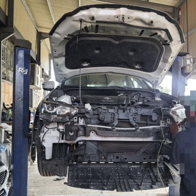 Voiture au capot ouvert sur un pont élévateur dans un garage, une personne travaille sur l'avant, des pièces sont démontées.