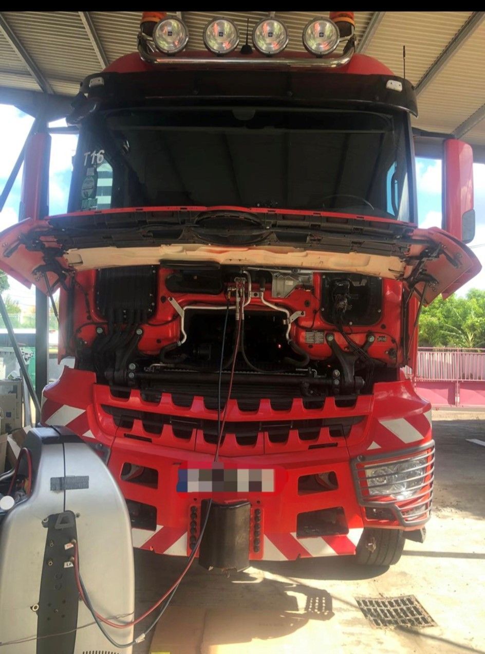 Camion semi-remorque rouge, capot ouvert pour réparation. Pièces du moteur noires visibles, bandes de signalisation blanches. Dans un garage.