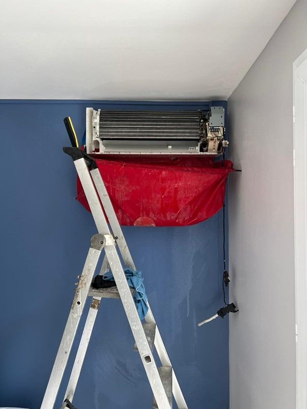 Échelle à côté du mur bleu, le climatiseur est en train d'être nettoyé, un sac rouge suspendu dessous.
