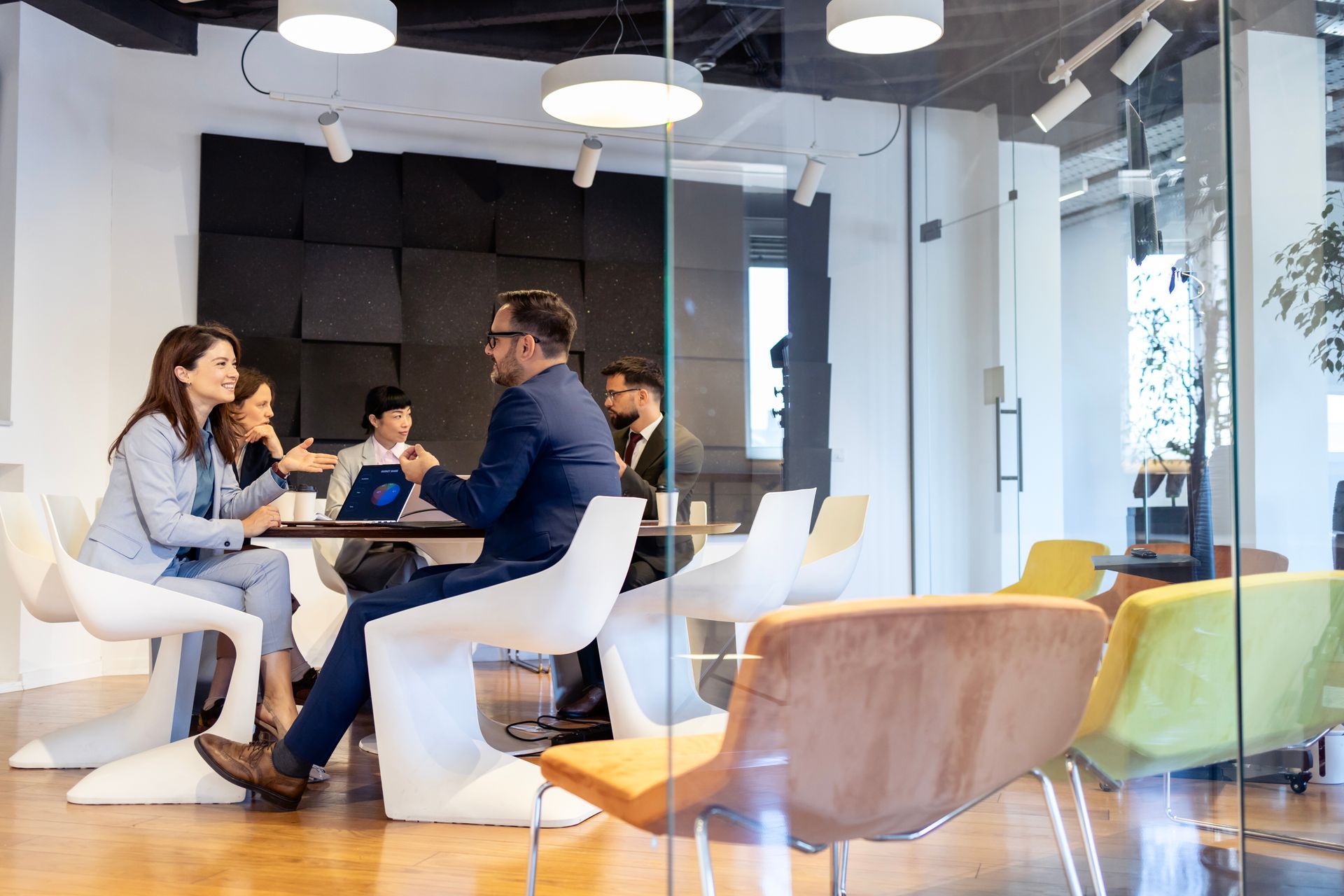 Menschen sitzen in einem modernen Büro an einem runden Tisch mit Laptops und Kaffee, hinter ihnen Glaswände.