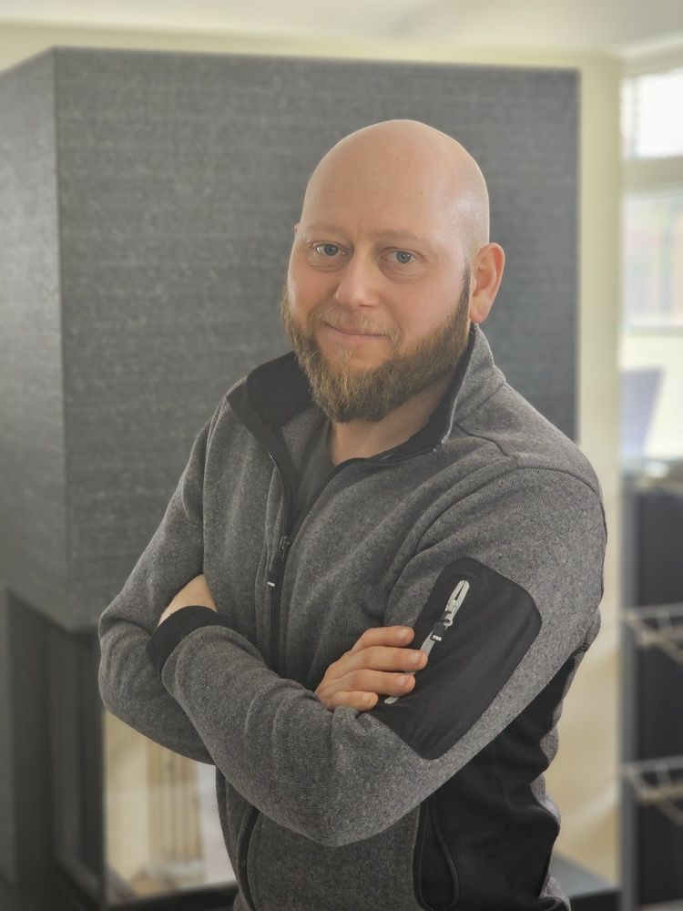 Ein kahlköpfiger Mann mit Bart steht mit verschränkten Armen vor einem Kamin.