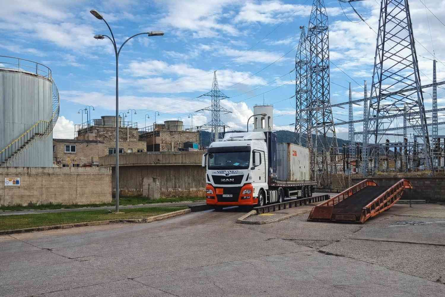 Tres camiones alineados bajo una grúa de carga en un puerto, con contenedores de envío apilados en el fondo.