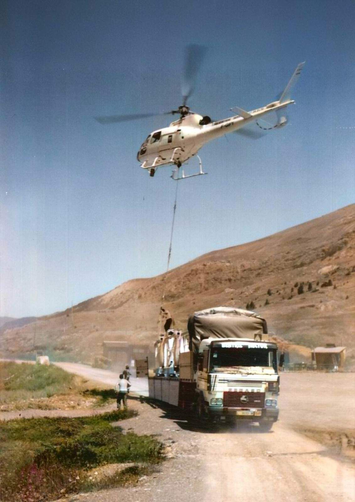 Helicóptero levantando carga de un camión en un camino de tierra en un entorno montañoso; cielo brillante.