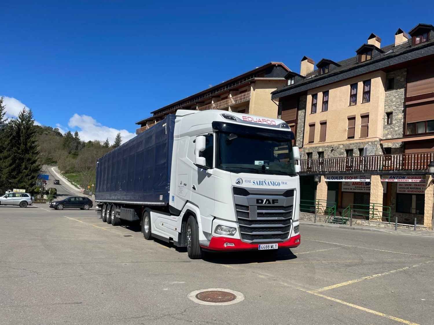 Un camión semirremolque viaja por una autopista, con montañas al fondo y un cielo azul claro.