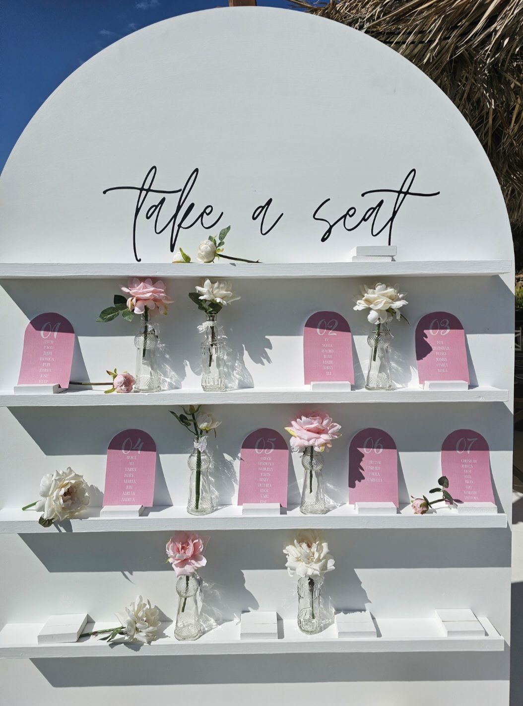 Una pared blanca con estantes llenos de jarrones con flores y un cartel que dice toma asiento.