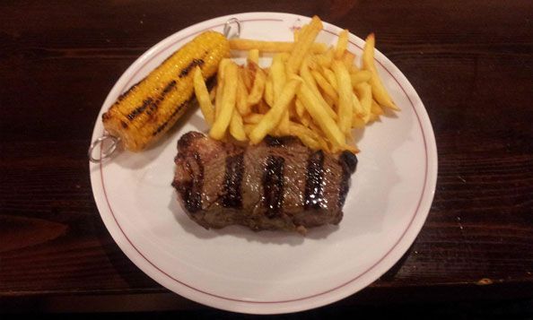 Un plato blanco cubierto con un filete, mazorcas de maíz y papas fritas.