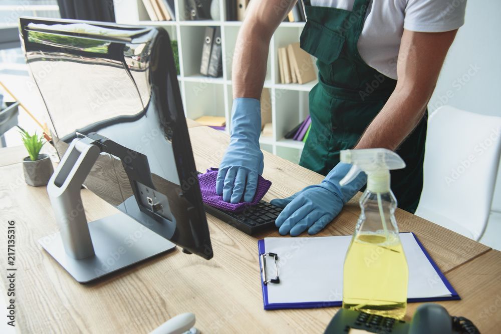 Eine Person reinigt mit Handschuhen, Sprühflasche, Monitor und Klemmbrett eine Tastatur an einem Schreibtisch.