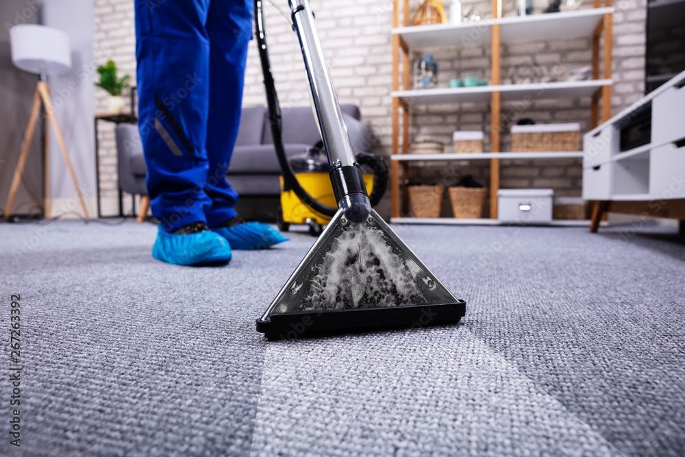 Teppichreinigungsdüse steht auf einem Teppich; daneben ist eine Person in blauer Hose und Stülpen in einem Wohnzimmer.
