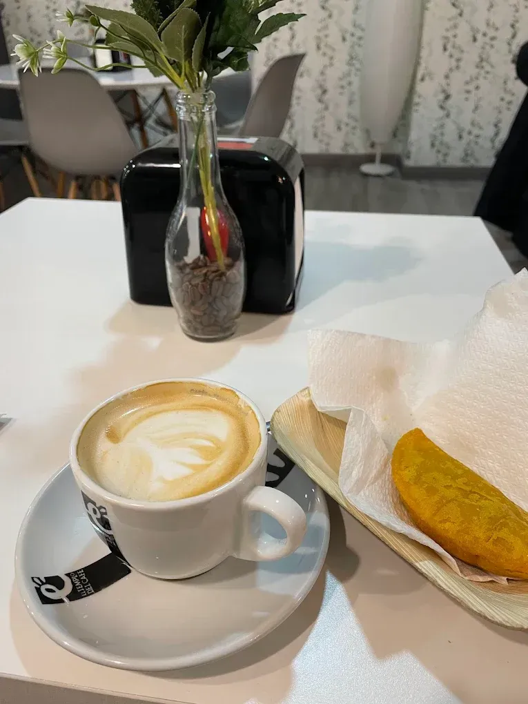 Capuchino en taza blanca, arepa en plato, jarrón con flores sobre mesa blanca.