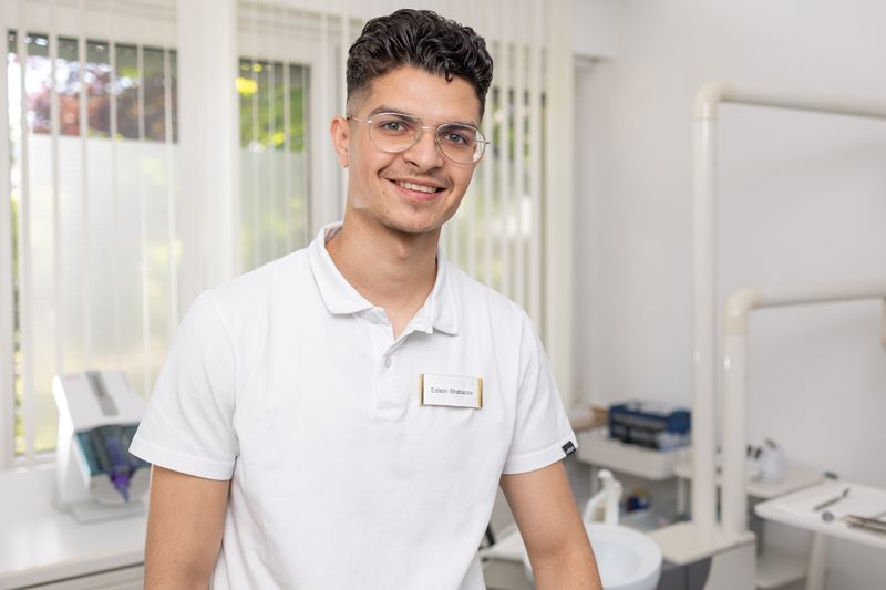 Ein junger Mann in einem weißen Poloshirt steht vor einer weißen Wand.