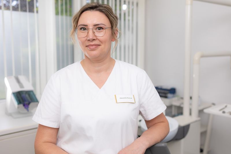 Eine Frau mit Brille und weißem Kasack steht vor einer weißen Wand.