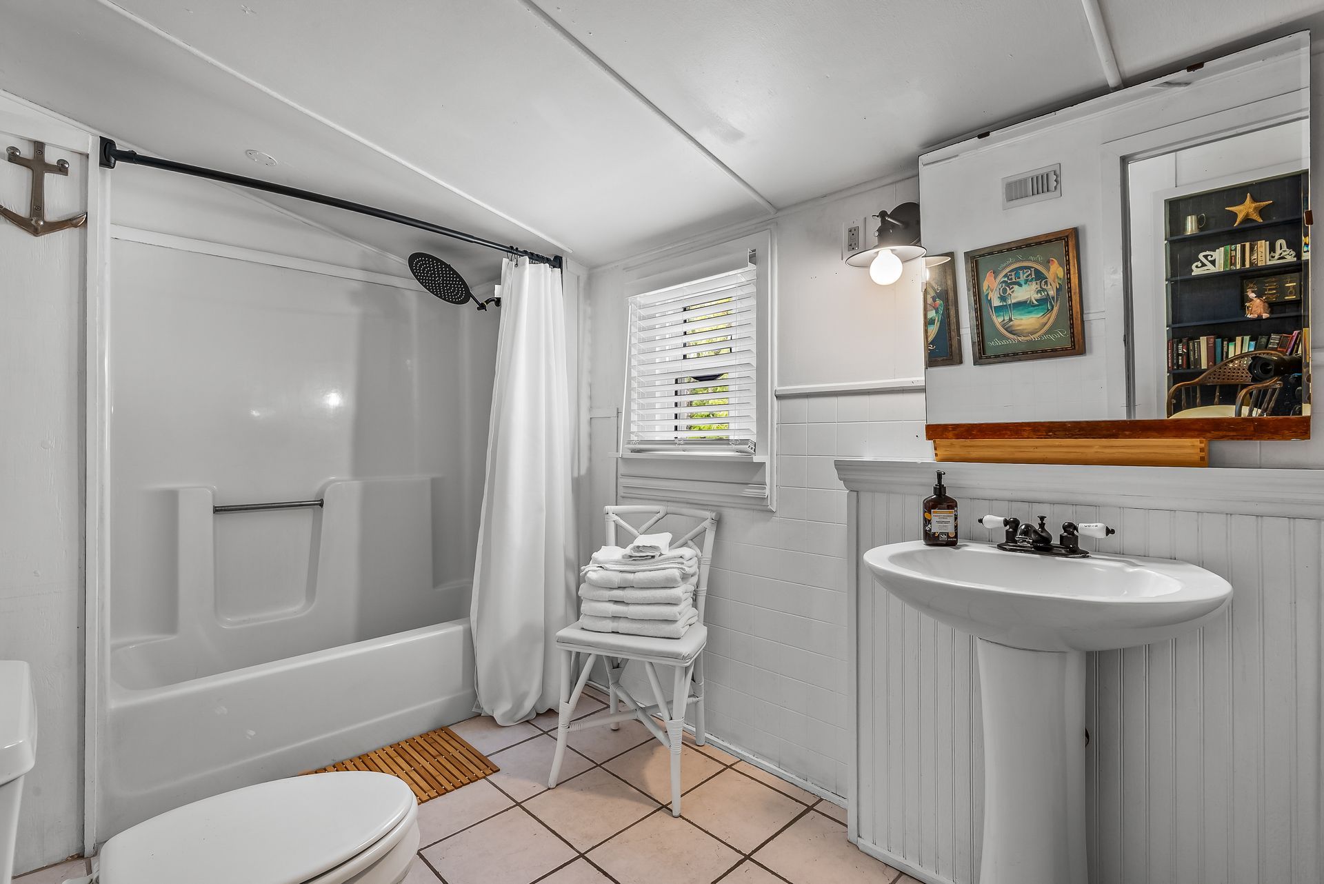 Bright bathroom with white tub, tiled floor, pedestal sink, mirror, and small window seat