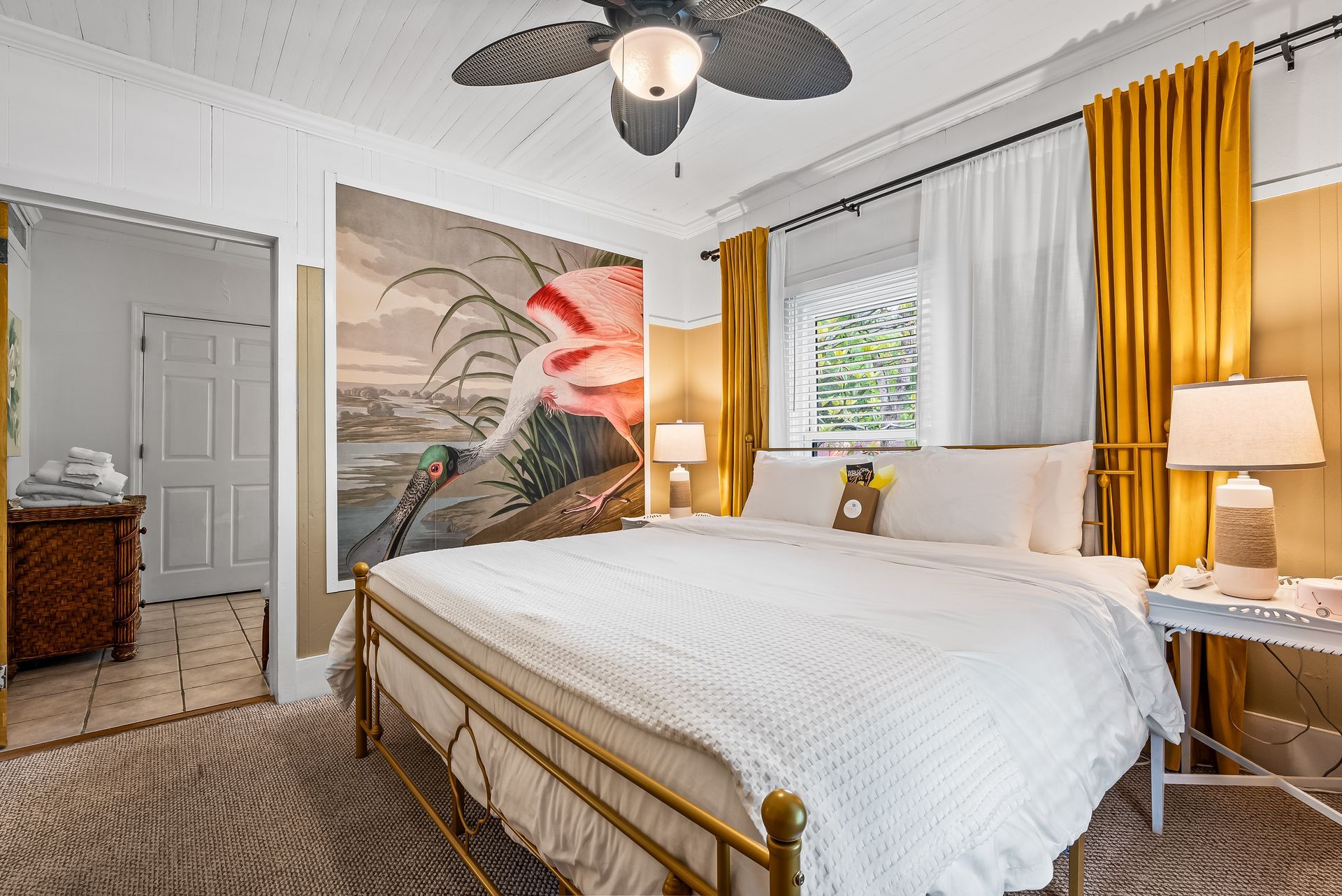 Bright Magnolia bedroom with white bedding, gold accents, mural art, and yellow curtains beside a window and lamp