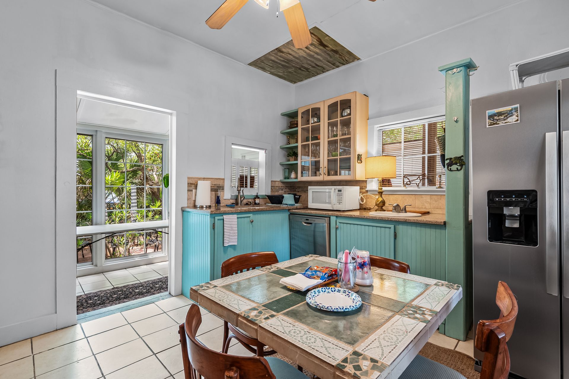 Community kitchen with turquoise cabinets, tiled floor, dining table, new fridge and open patio door to greenery.