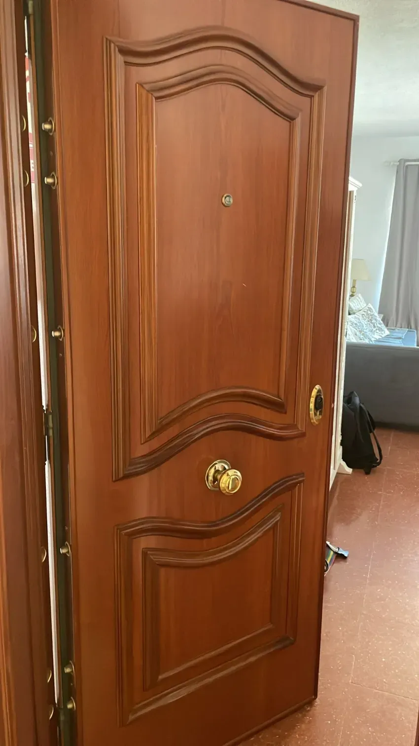 En un dormitorio, se abre una puerta de madera con pomo de latón, dejando ver parcialmente una cama y una cortina en el interior.