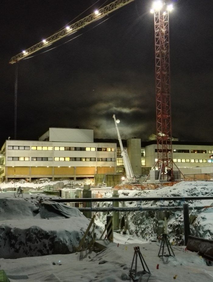 Yöllinen rakennustyömaa, jossa on korkea nosturi, valaistuja rakennuksia ja lumen peittämä maa