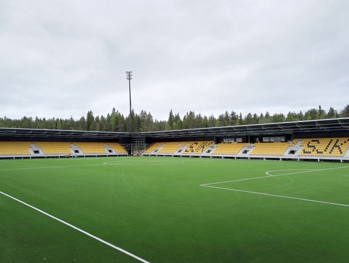 Tyhjä jalkapallostadion vihreällä kentällä, kelta-mustilla katsomoilla ja pilvisellä taivaalla