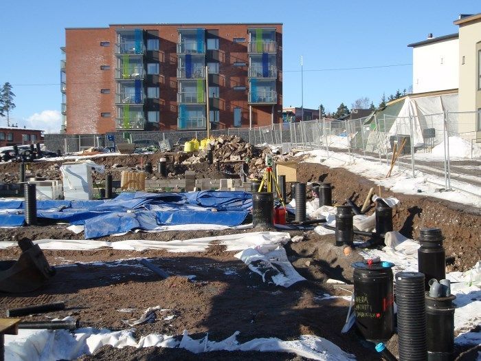 Rakennustyömaa, jossa on kaivettu maa, sinisiä pressuja ja taustalla tiilikerrostalo