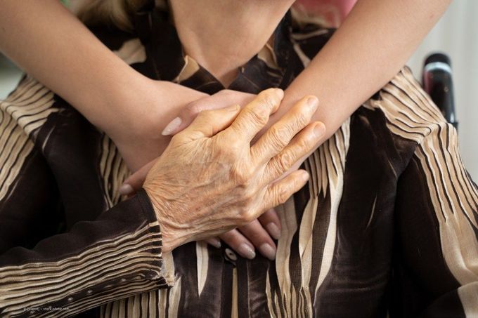Oberkörper einer Seniorin, der von hinten von zwei Frauenarmen umarmt wird, die rechte Hand der Seniorin liegt auf den überkreuzten Händen der Frau hinter ihr