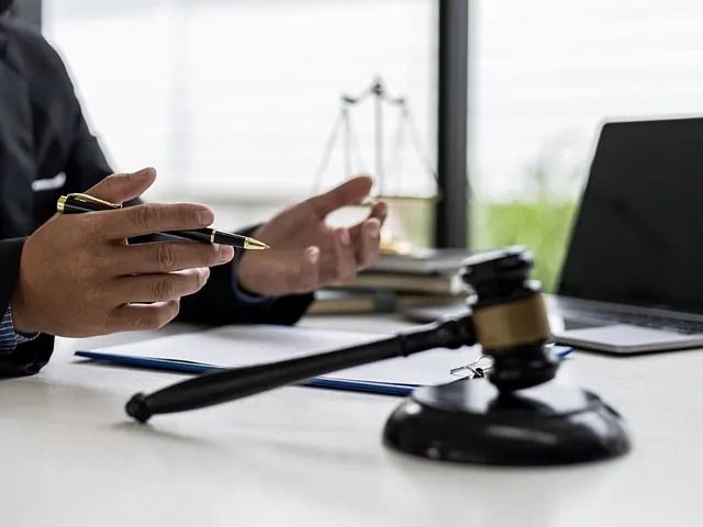 Una persona con traje gesticula con un bolígrafo, un mazo sobre una mesa con una computadora portátil y una balanza de la justicia en el fondo.