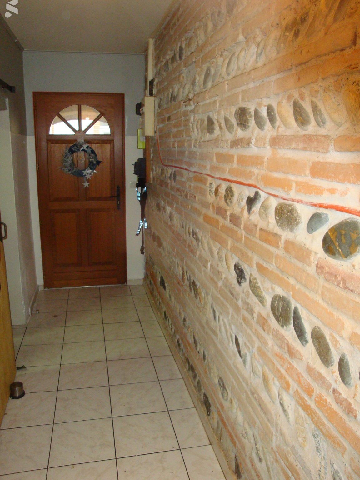 Couloir étroit avec mur de briques orné de pierres incrustées ; porte en bois au fond, sol carrelé.