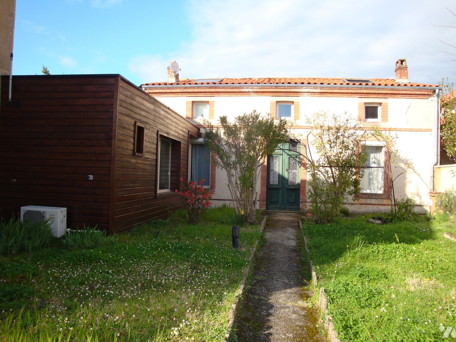 Vue arrière d'une maison beige avec une structure en bois marron attenante, une pelouse envahie par les herbes hautes et un chemin.